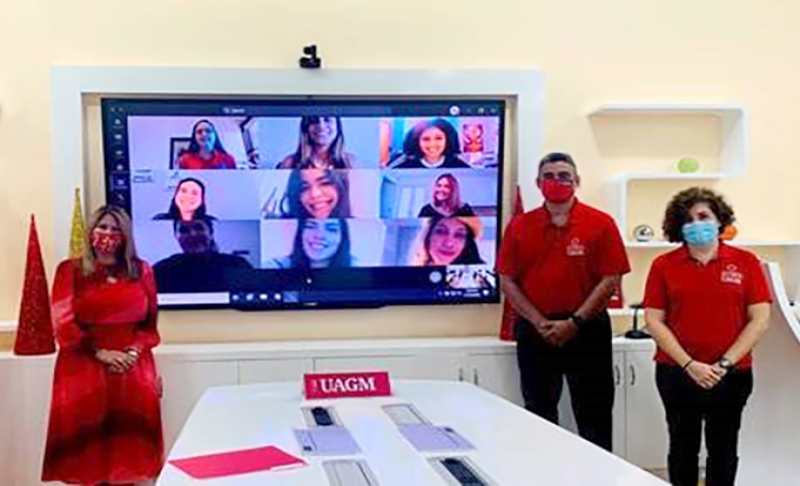 Tres ejecutivos de UAGM parados frente a un televisor donde se proyectan un grupo de estudiantes virtual
